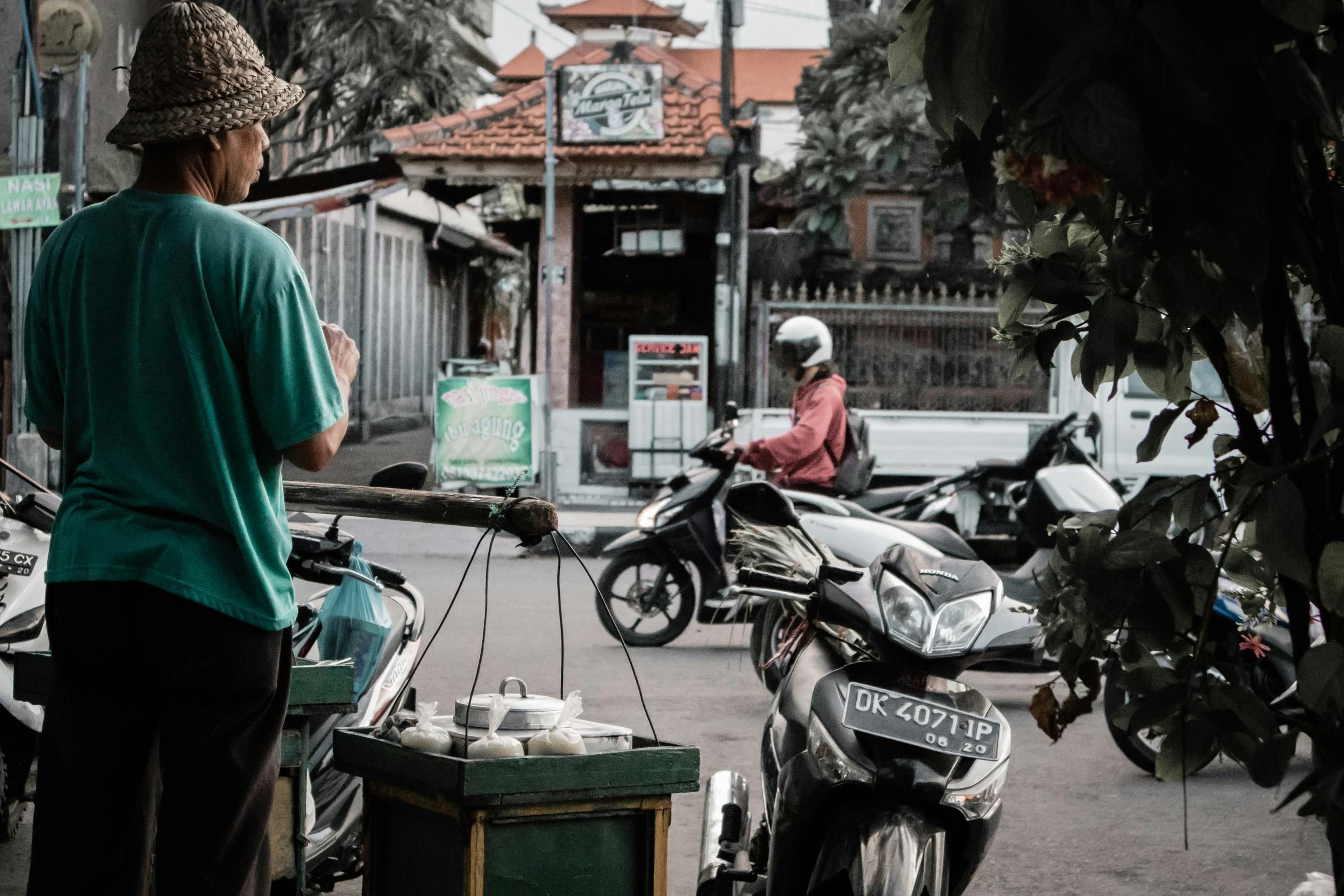Bali — A street vendor in Marga, Bali, Indonesia, sells goods to passing motorcyclists. Busy street life scene.