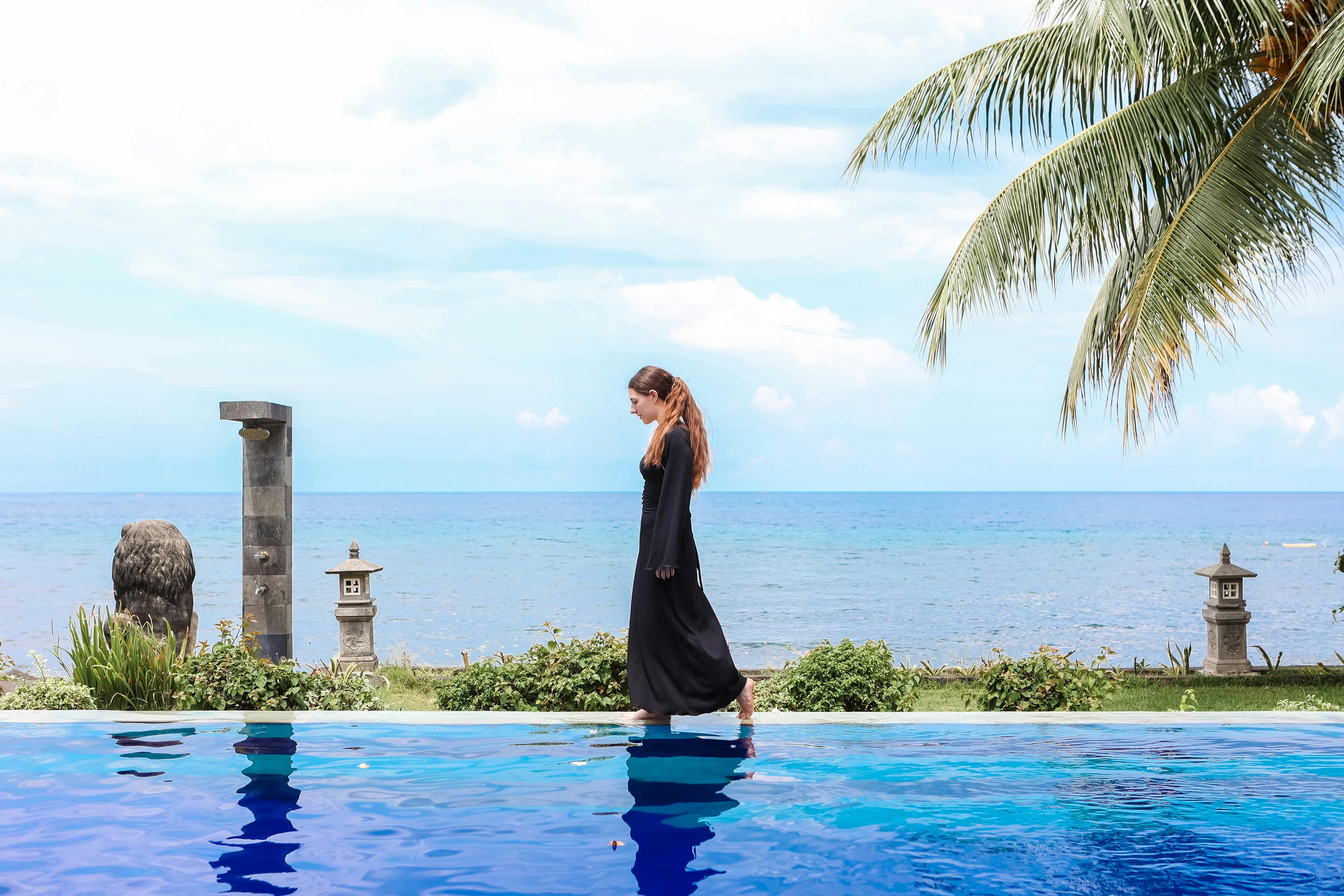 Bali — Serene scene of a woman walking by a pool in Bali with ocean view, palm tree, and sunny sky.