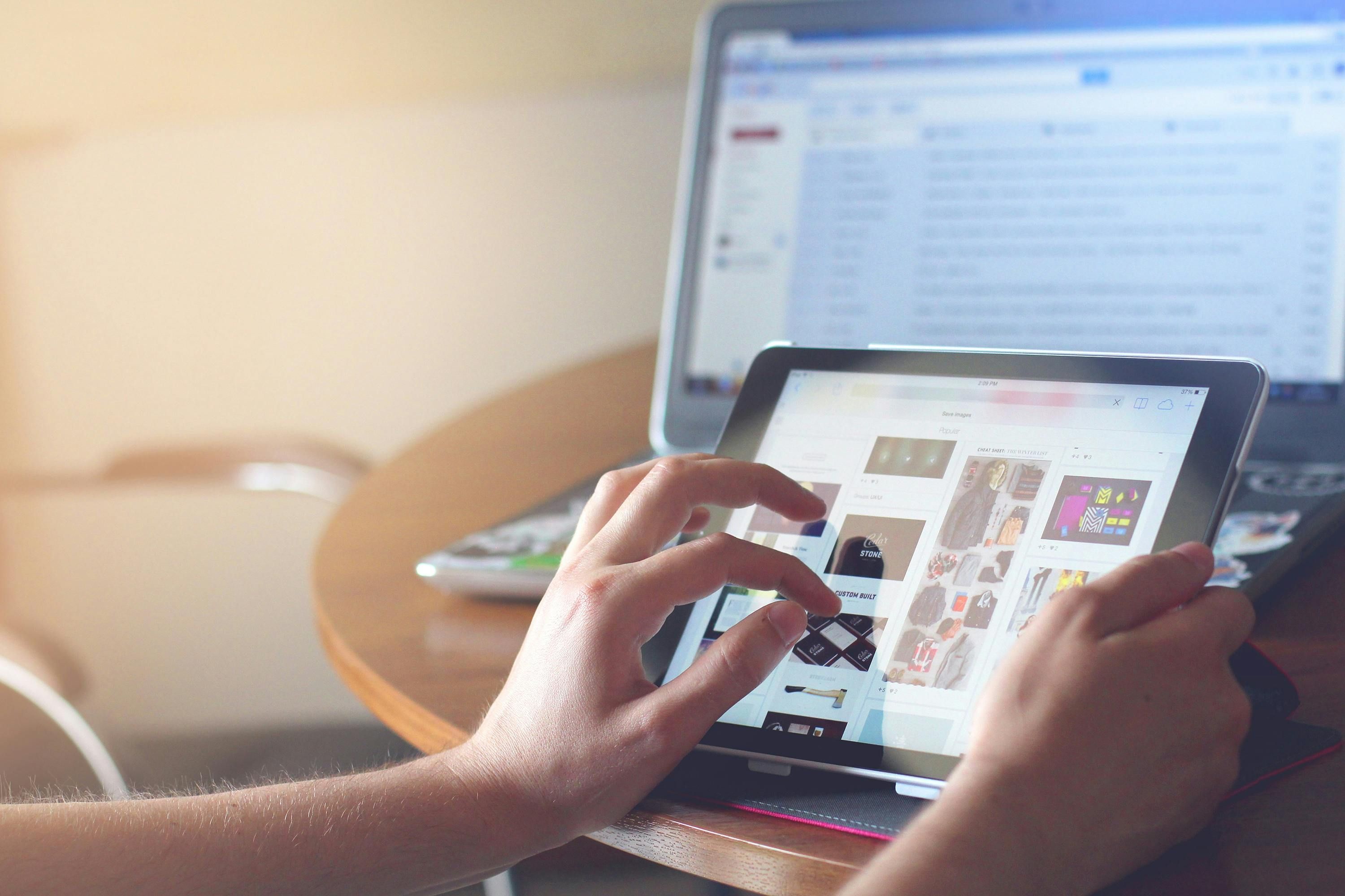 Bali — Hands using a tablet beside a laptop in a home office setting, showcasing online browsing.