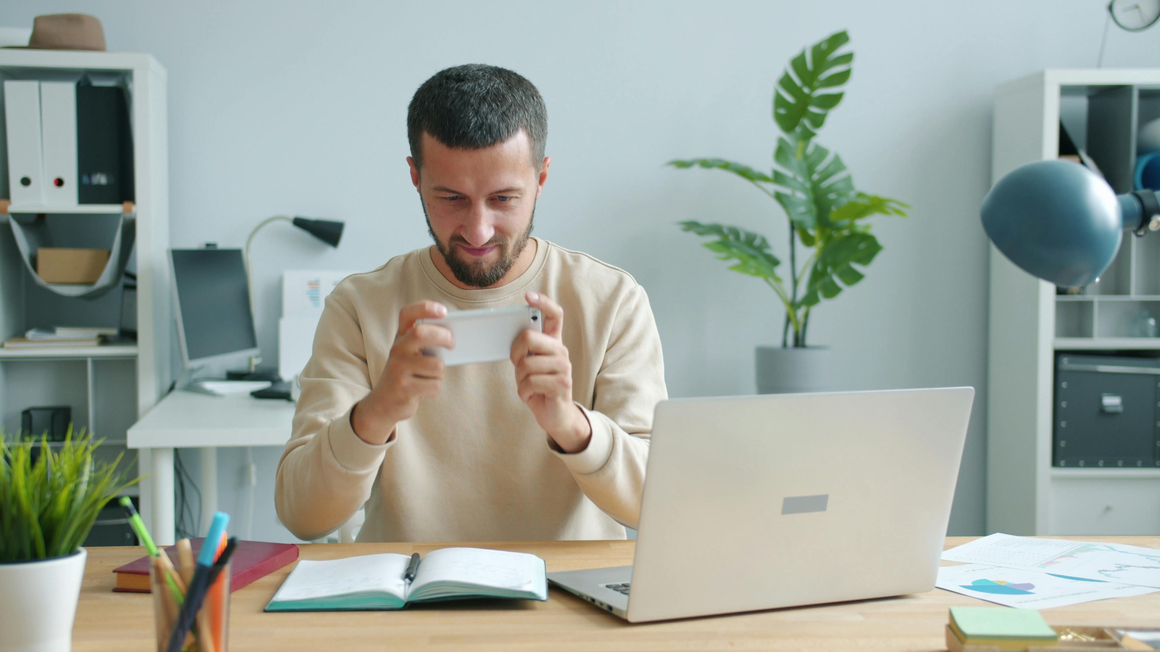 Bali — Focused man using smartphone at a desk with laptop in a modern office setting.
