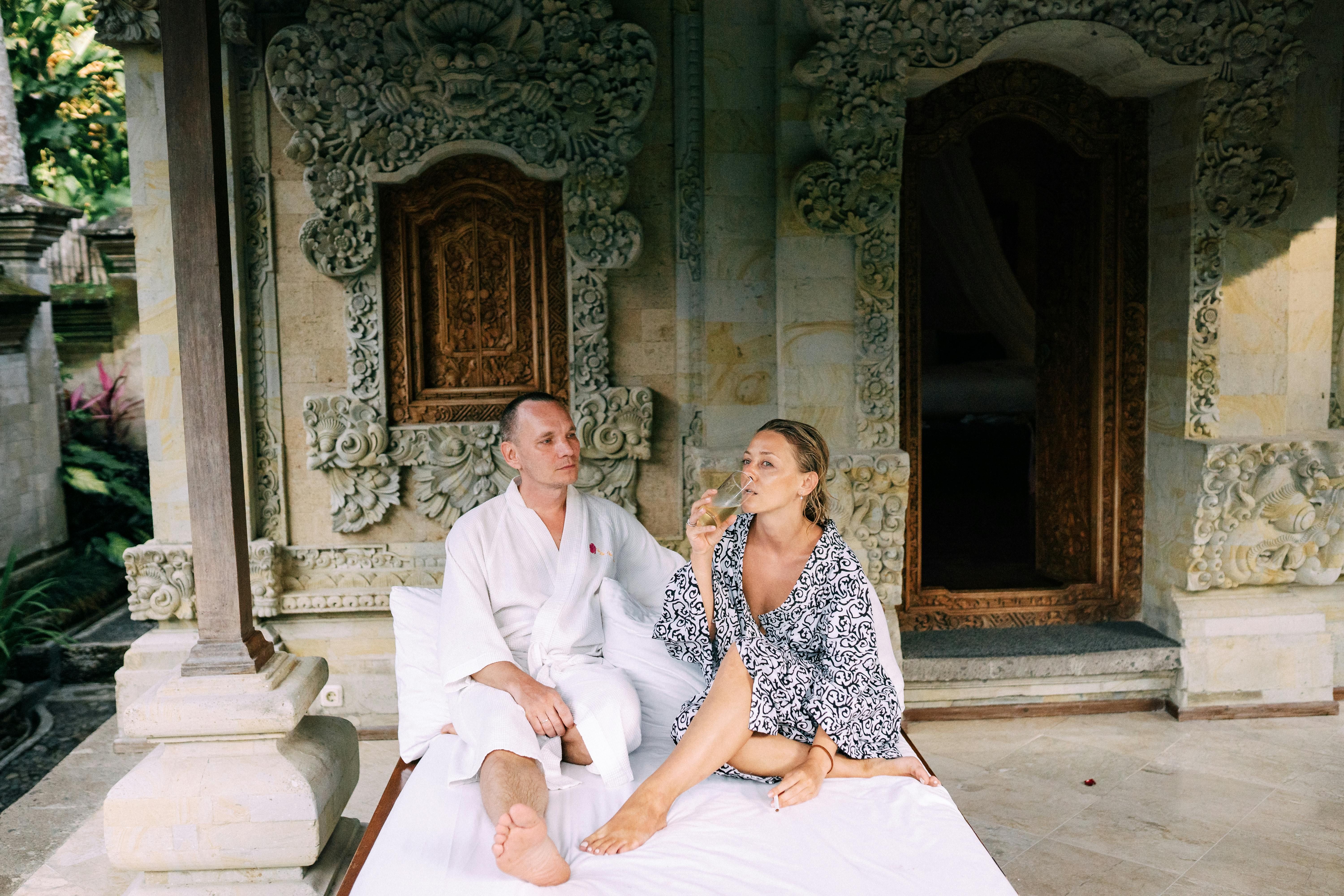 Bali — A couple in robes enjoy a relaxing moment outside a Balinese villa with ornate carvings.
