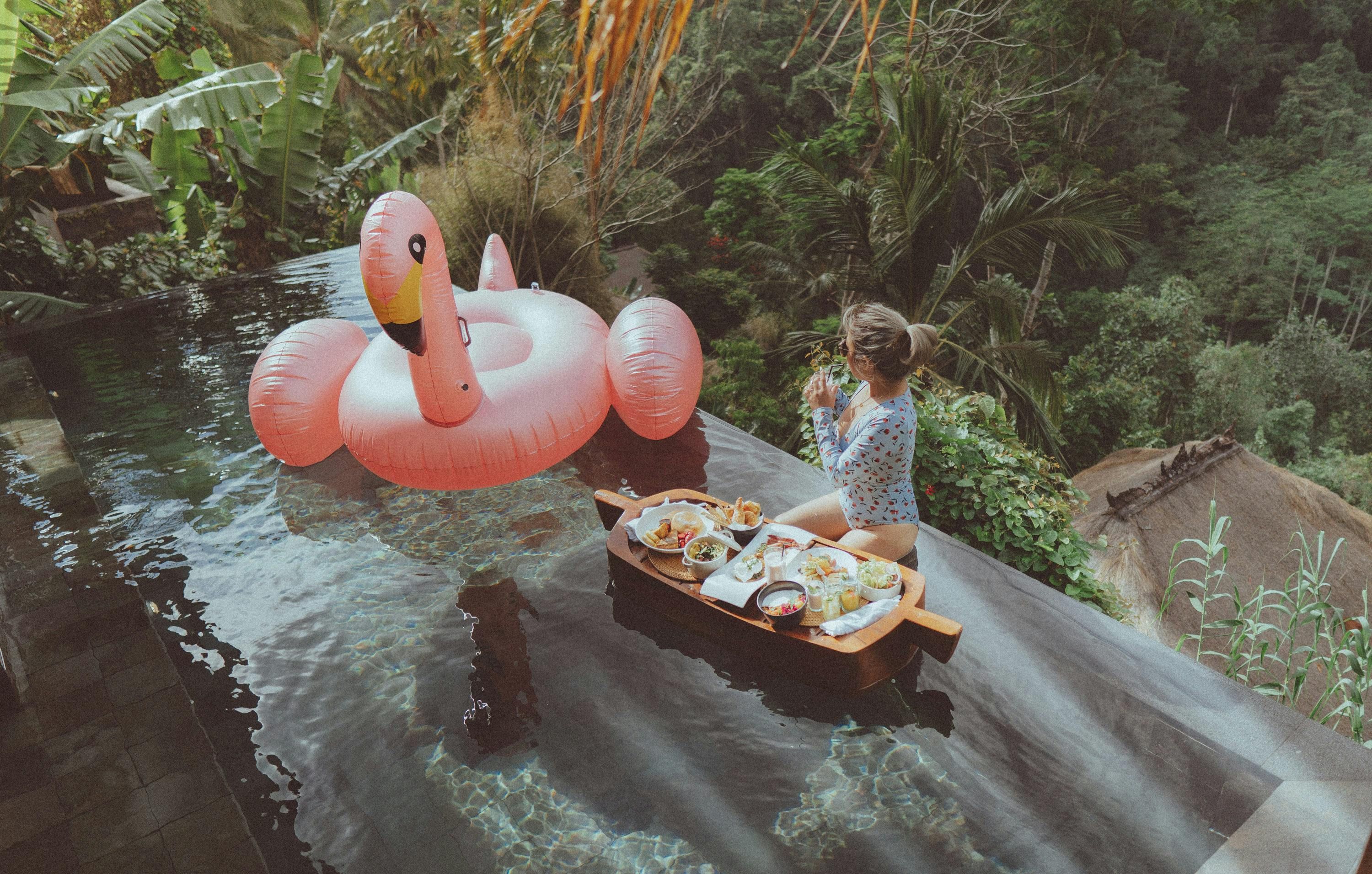Bali — Woman enjoying breakfast in an infinity pool with a flamingo float at a Bali resort. Tropical serenity and relaxation.