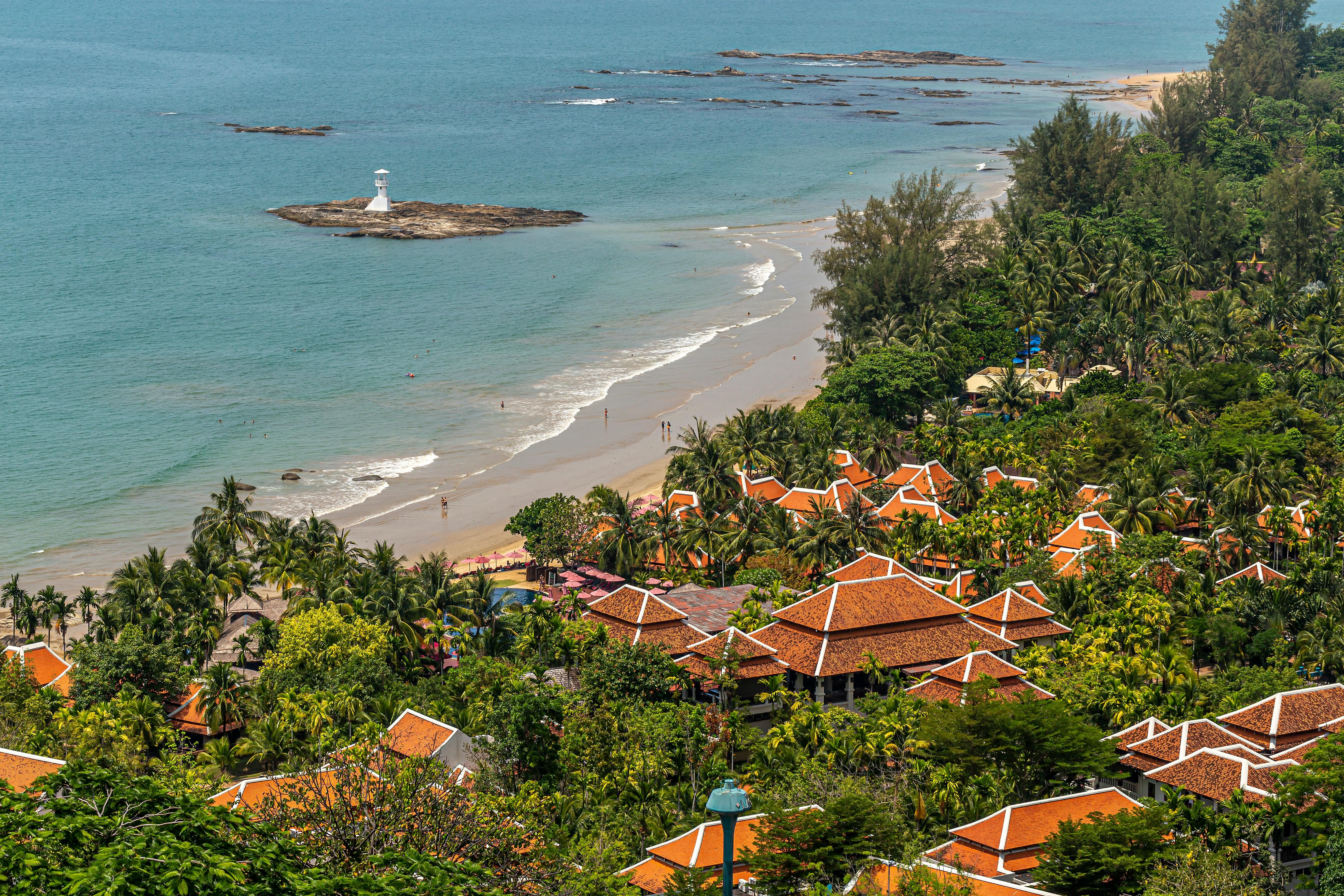 Thailand — A picturesque beachfront village with terracotta roofs, palm trees, and a distant lighthouse.